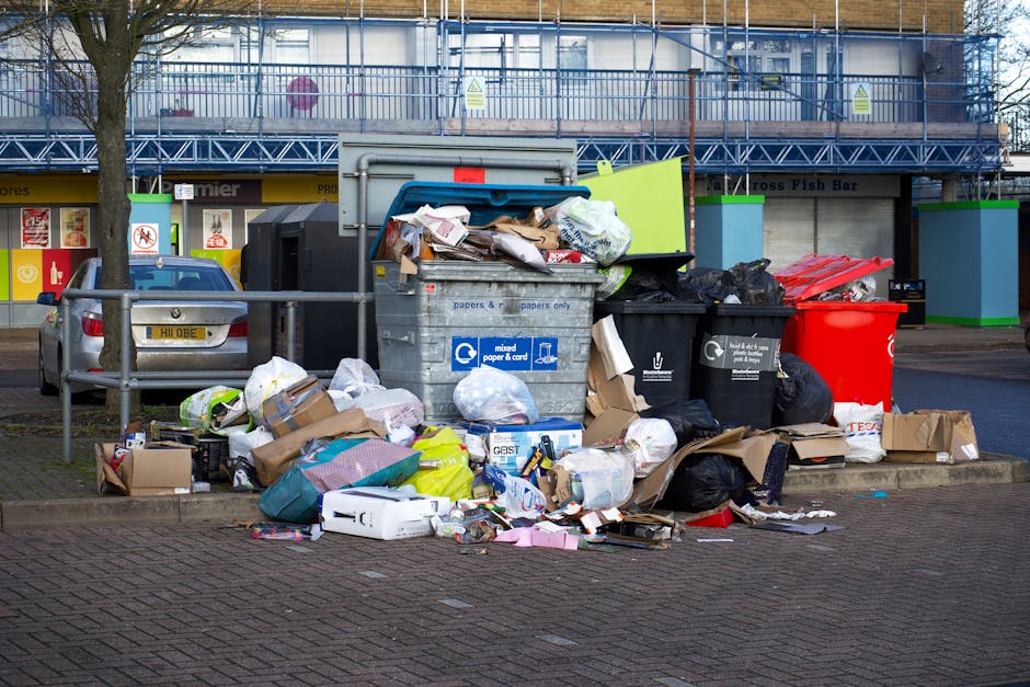 A street scene in an urban area featuring a row of tall, multi-story buildings with varied architectural styles, including brick facades, arched windows, and decorative elements. In the foreground, a waste collection vehicle, labeled with the logo of Waste Disposal Clapham, is parked on the right side of the street, ready for rubbish collection. The vehicle is a modern, light grey truck with a compact design, situated on a paved road with black bollards and several bicycle racks aligned along the sidewalk. The street is illuminated by natural daylight with overcast weather, creating a neutral, soft light that highlights the textures of the buildings and urban environment. To the left, a stone entrance gate with metal gates and a small plaque is partially visible, indicating access to a private or council-managed property. The surrounding environment suggests a typical city setting with adjacent buildings closely packed, windows reflecting the muted sky, and a general atmosphere suited to independent waste collection or private rubbish removal services provided by Waste Disposal Clapham, aligned with alternative waste handling approaches common in urban settings.