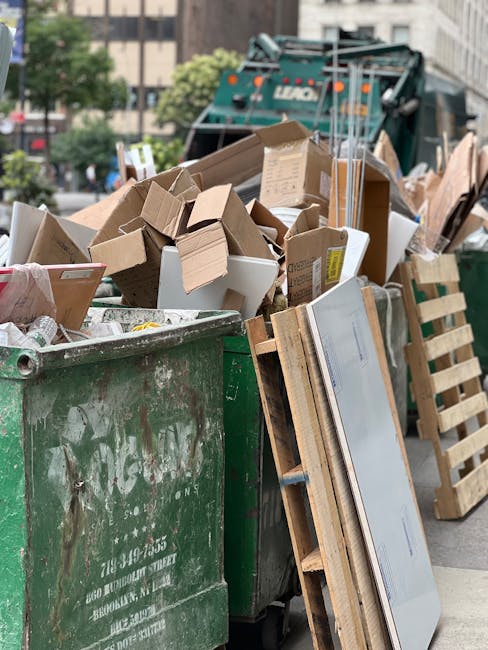 A large green waste disposal skip filled with various discarded cardboard boxes of different sizes, some flattened and some still in their original shapes, along with a few white and brown paper packaging materials. The boxes show visible corrugated textures and are mostly brown, with some partially covered with labels or packaging tape. Leaning against the side of the skip is a wooden pallet with slatted planks, and nearby, there are small stacks of flat cardboard sheets. In the background, a green waste collection vehicle is partially visible, with blurred urban buildings and trees surrounding the scene, indicating an outside street setting during daylight hours. The scene captures a typical private waste collection operation, representing rubbish removal services, with natural lighting highlighting the textures and colors of the materials being prepared for disposal, consistent with independent bulk waste clearance activities that Waste Disposal Clapham might undertake for clients in the area.