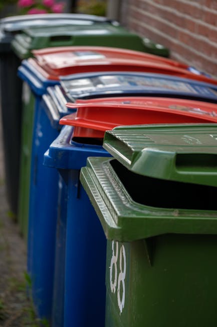 A row of large wheelie bins aligned along a sidewalk against a brick wall, with green, blue, red, and black lids, each lid with a textured surface. The bins are made of plastic and have a smooth finish, with some showing signage or markings on the front. The bins are positioned in close proximity, with the green bin in the foreground and others extending into the background. The environment appears to be an outdoor urban or residential area, with natural daylight casting soft shadows. These bins are typically used for waste collection and disposal, reflecting alternative or private rubbish removal services, as provided by Waste Disposal Clapham, often involved in on-site clearance or alternative waste handling methods, supporting local rubbish removal needs.