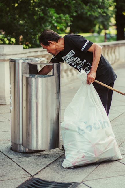A middle-aged man with dark hair, wearing a black T-shirt with patterned text and dark trousers, is engaged in rubbish disposal at an outdoor location. He is bending slightly forward, placing waste into a modern, stainless steel public bin with multiple separate compartments, situated on a paved walkway. The man holds a large white trash bag in his left hand, which appears to be filled with rubbish, and grips a wooden broom or litter picker with his right hand, used to assist in cleaning or collecting waste. The surrounding environment features lush trees with green foliage and a stone balustrade in the background, indicating the scene takes place in a park or public outdoor area. The lighting suggests daytime, with natural sunlight illuminating the scene softly, contributing to a clean and orderly atmosphere. This image exemplifies active participation in waste management or rubbish clearance, aligning with private or independent rubbish collection services such as those offered by Waste Disposal Clapham, which specialise in rubbish removal outside of local authority collections.