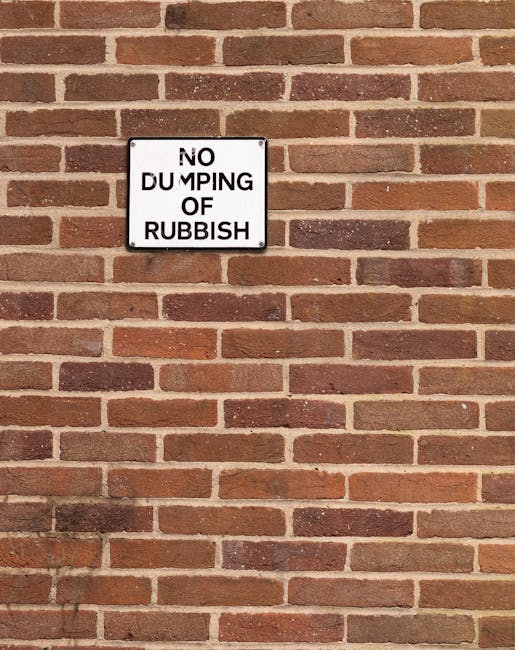 A close-up view of a brick wall painted in shades of red, brown, and tan, with a rectangular white metal sign mounted near the upper left corner. The sign displays the message 'NO DUMPING OF RUBBISH' in black, bold, capitalized letters, with the word 'DUMPING' partially obscured by wear or damage. The brick wall has a textured surface with visible mortar lines, extending horizontally across the image. The lighting is natural, casting soft shadows, and the overall scene emphasizes a quiet urban or residential environment where private waste management or rubbish clearance services, such as those provided by Waste Disposal Clapham, may be relevant, especially in the context of promoting alternative rubbish collection options in Clapham.
