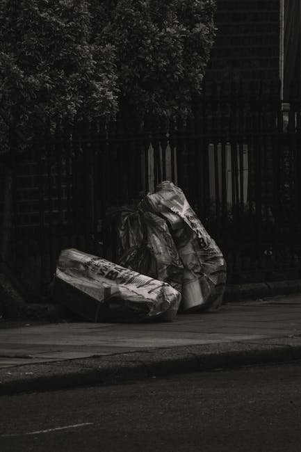 A middle-aged man with dark hair, wearing a black T-shirt with patterned text and dark trousers, is engaged in rubbish disposal at an outdoor location. He is bending slightly forward, placing waste into a modern, stainless steel public bin with multiple separate compartments, situated on a paved walkway. The man holds a large white trash bag in his left hand, which appears to be filled with rubbish, and grips a wooden broom or litter picker with his right hand, used to assist in cleaning or collecting waste. The surrounding environment features lush trees with green foliage and a stone balustrade in the background, indicating the scene takes place in a park or public outdoor area. The lighting suggests daytime, with natural sunlight illuminating the scene softly, contributing to a clean and orderly atmosphere. This image exemplifies active participation in waste management or rubbish clearance, aligning with private or independent rubbish collection services such as those offered by Waste Disposal Clapham, which specialise in rubbish removal outside of local authority collections.